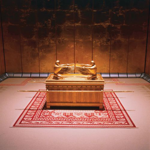 A replica of the ark of the covenant, covered in gold, standing on a woven red and white rug.
