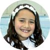 A young girl in a white dress sits outside.  It looks to be that this is her baptism photo.  She has dark long curled hair with a white headband.  She is sitting on the edge of a fountain.