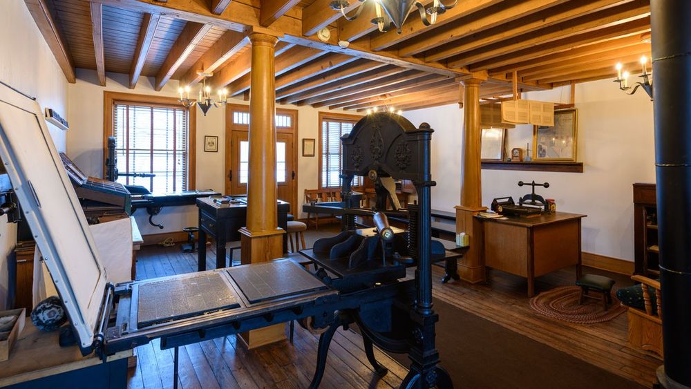 Large black press in a press room with newspaper type set and platen raised.