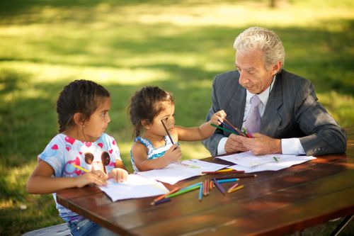 A grandfather sits outside at a picnic table and colors with his two young granddaughters.