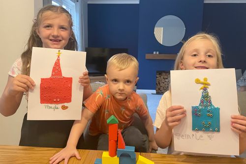 Two girls holding up drawings of a temple and a boy with a block model of a temple