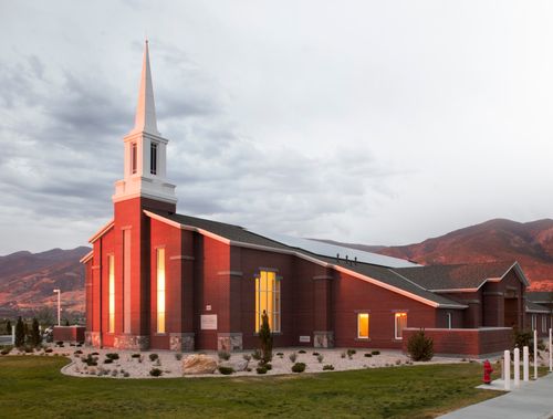 The sun shining on the front of a red-brick chapel with a white steeple surrounded by green grass and mountains in the background.