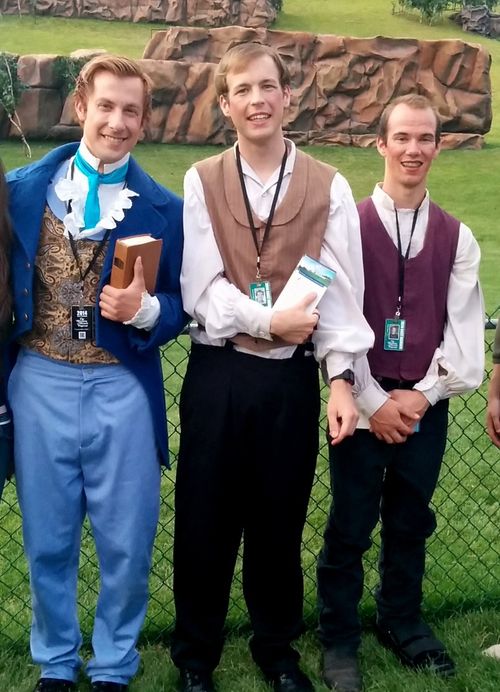 Three actors smiling and standing against a black wire fence with mountain props in the background at the Manti Pageant.