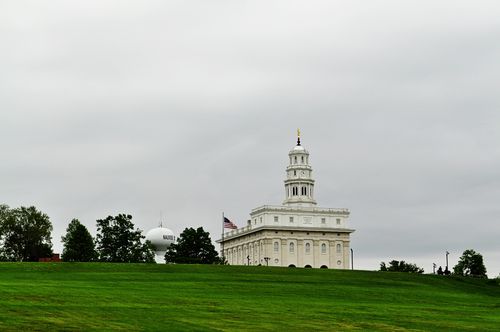 The Nauvoo Illinois Temple seen from the bottom of the hill, with a green lawn topped with dark green trees.
