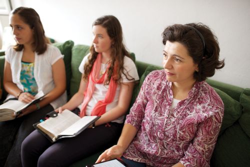 A woman and two teenagers sit on a green couch with their scriptures on their laps.