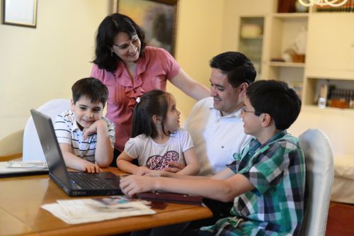 A father, mother, and three children from Argentina gather around a laptop sitting on a table as they work on family history together.
