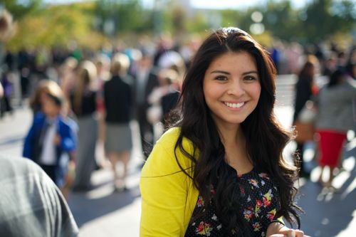 A woman with long dark hair and a bright yellow cardigan, standing outside of the Conference Center and smiling.