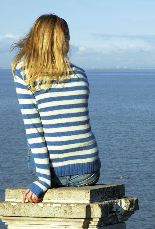 A young woman sitting on a concrete wall looking out over the ocean. photo of a blond girl looking out toward the sea