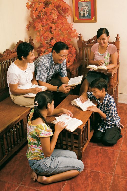 An Asian family gathered for scripture study.  They are sitting on sofas and the floor.