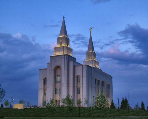 The Kansas City Missouri Temple, with the lights on in the evening and the green lawns surrounded by a black fence.
