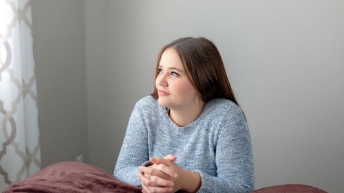 Young adult woman kneeling by bed pondering, listening