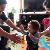 A father and mother play with their young baby boy. He is learning how to walk and his parents are trying to help him make those first steps.