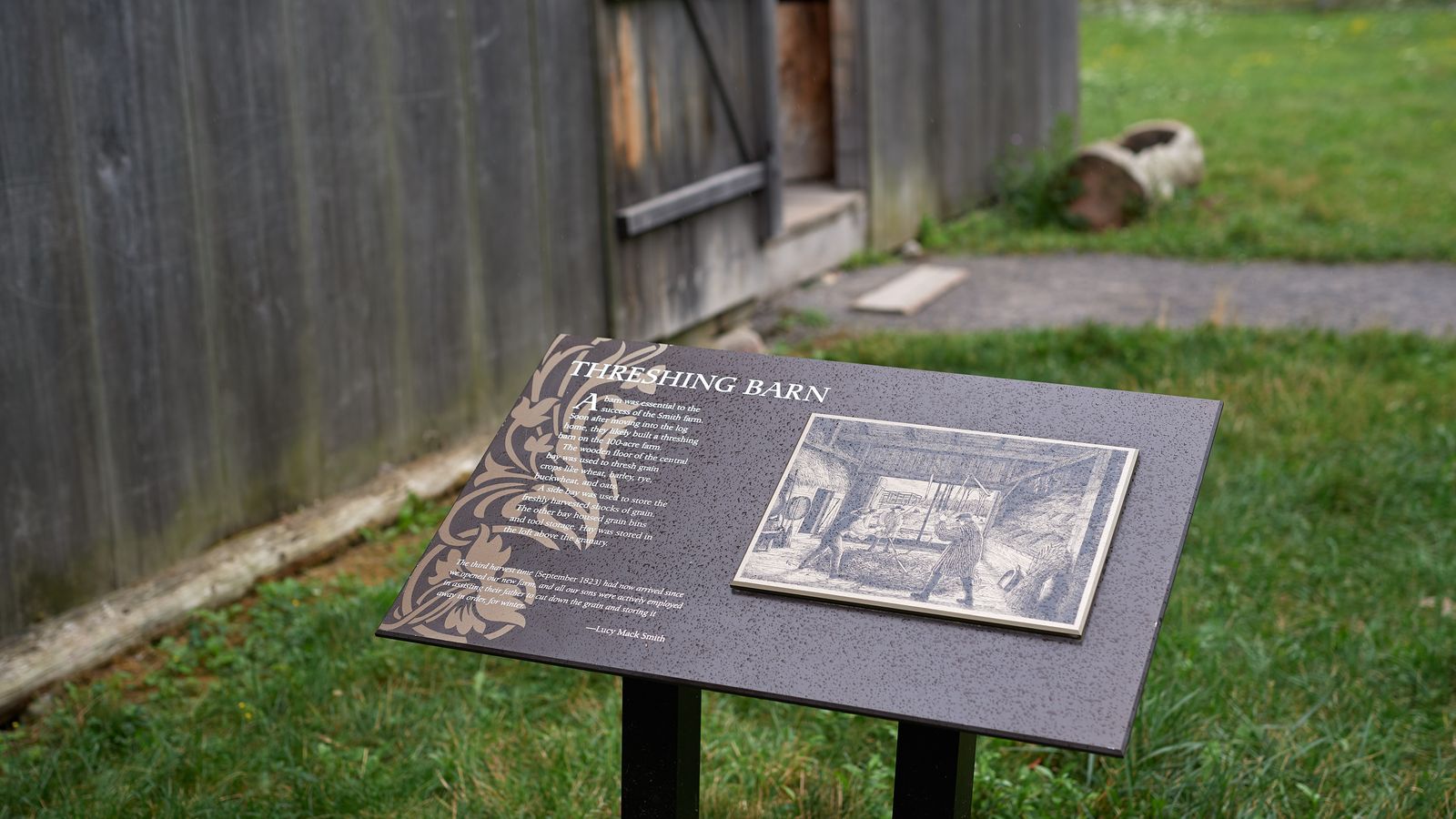 Various plaques around Palmyra and the Sacred Grove. This plaque says, 'Threshing barn.' It is surrounded by farmland and a rough wooden building in the background.