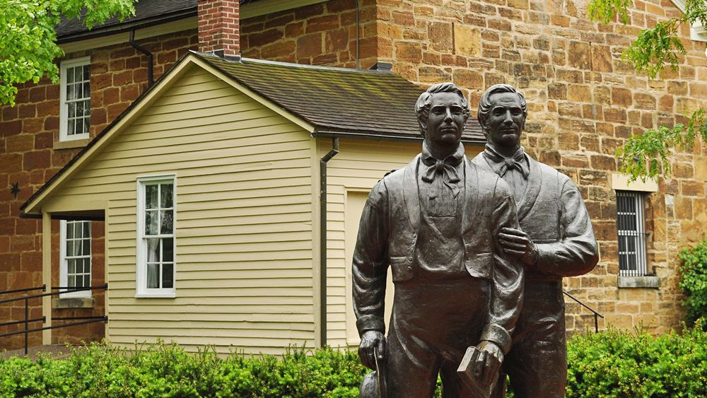 A bronze statue of two men stands in front of a clapboard house and a large brick two-story jail.