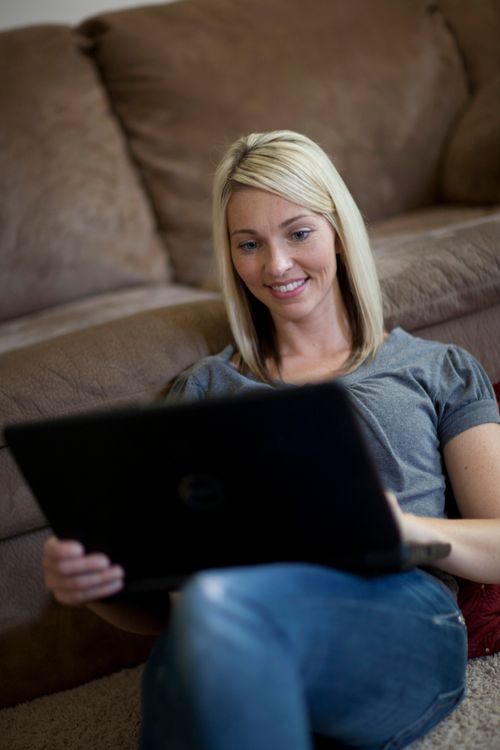 A woman sits on the floor, leaning against a couch, while looking at the laptop on her lap.