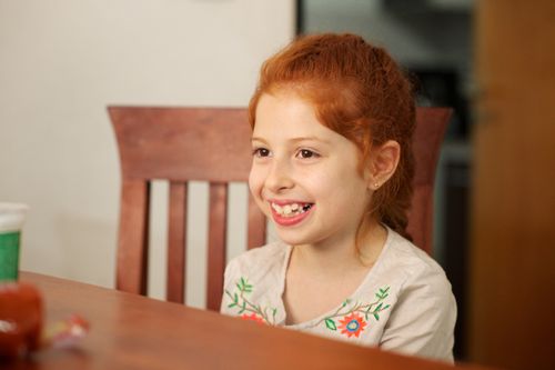 A young girl with red hair in a French braid, smiling and sitting at a dining-room table.