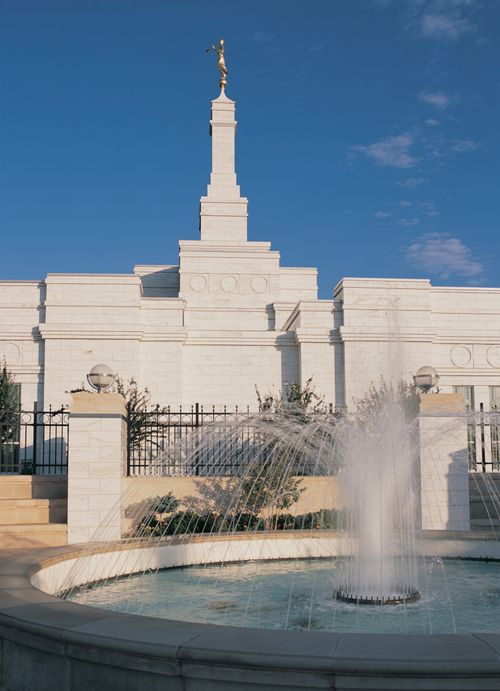 A water fountain on the grounds of the Oklahoma City Oklahoma Temple on a sunny day, with the temple seen on the left side and a blue sky overhead.