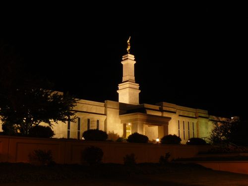 The front entrance side of the Monticello Utah Temple lit up at nighttime, with scenery of bushes and trees and with the angel Moroni atop the spire.