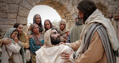 A young man wrapped in burial clothe is sitting up in a gurney. Jesus has his hand on his shoulder, his disciples can be seen in the background.