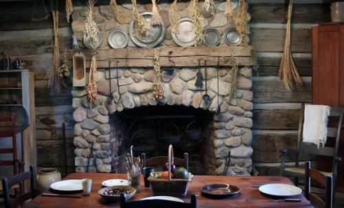 A fireplace, a dining table with plates and a fruit basket, and dried plants hanging from the ceiling in the Peter Whitmer cabin in Fayette, New York.