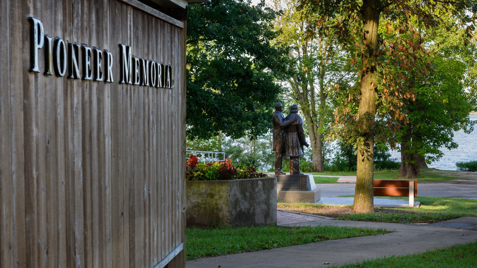 The side of a wooden building with a sign that reads 'Pioneer Memorial.' In the foreground are trees and the back of a bronze statue of Brigham Young and Joseph Smith