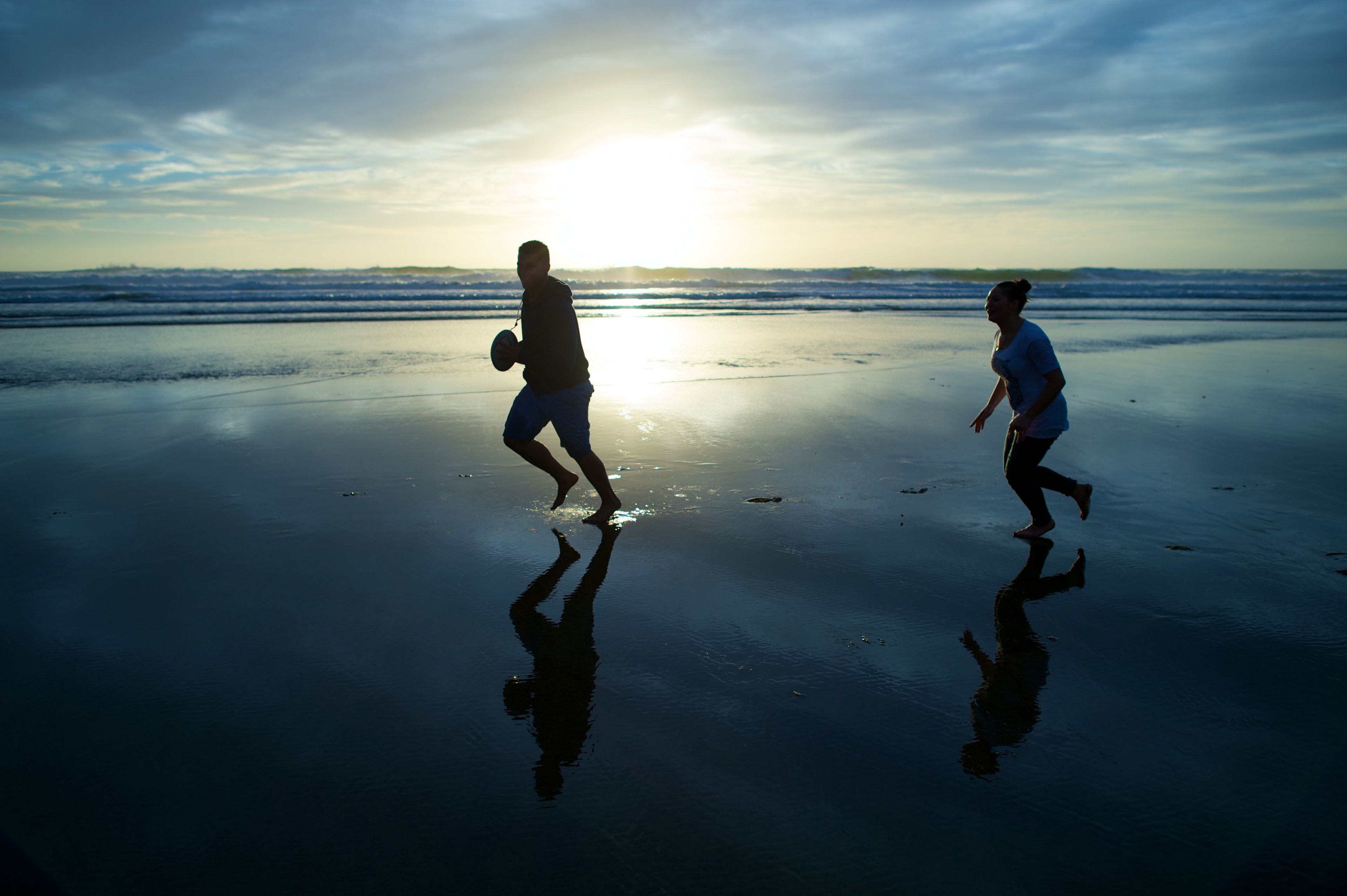 A young man and a young woman play with a football on the beach at sunset.