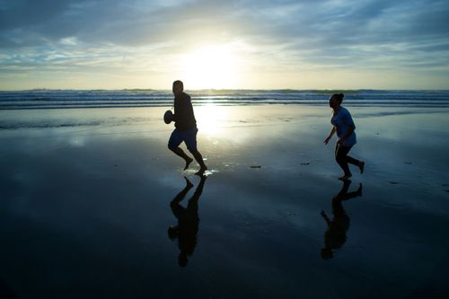 A young man runs through water on the beach carrying a football, with a young woman chasing him as the sun sets.