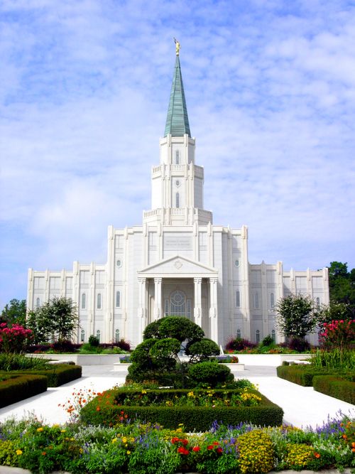 The front of the Houston Texas Temple on a sunny day, with flower beds and green trees forming a median on the path to the doors.