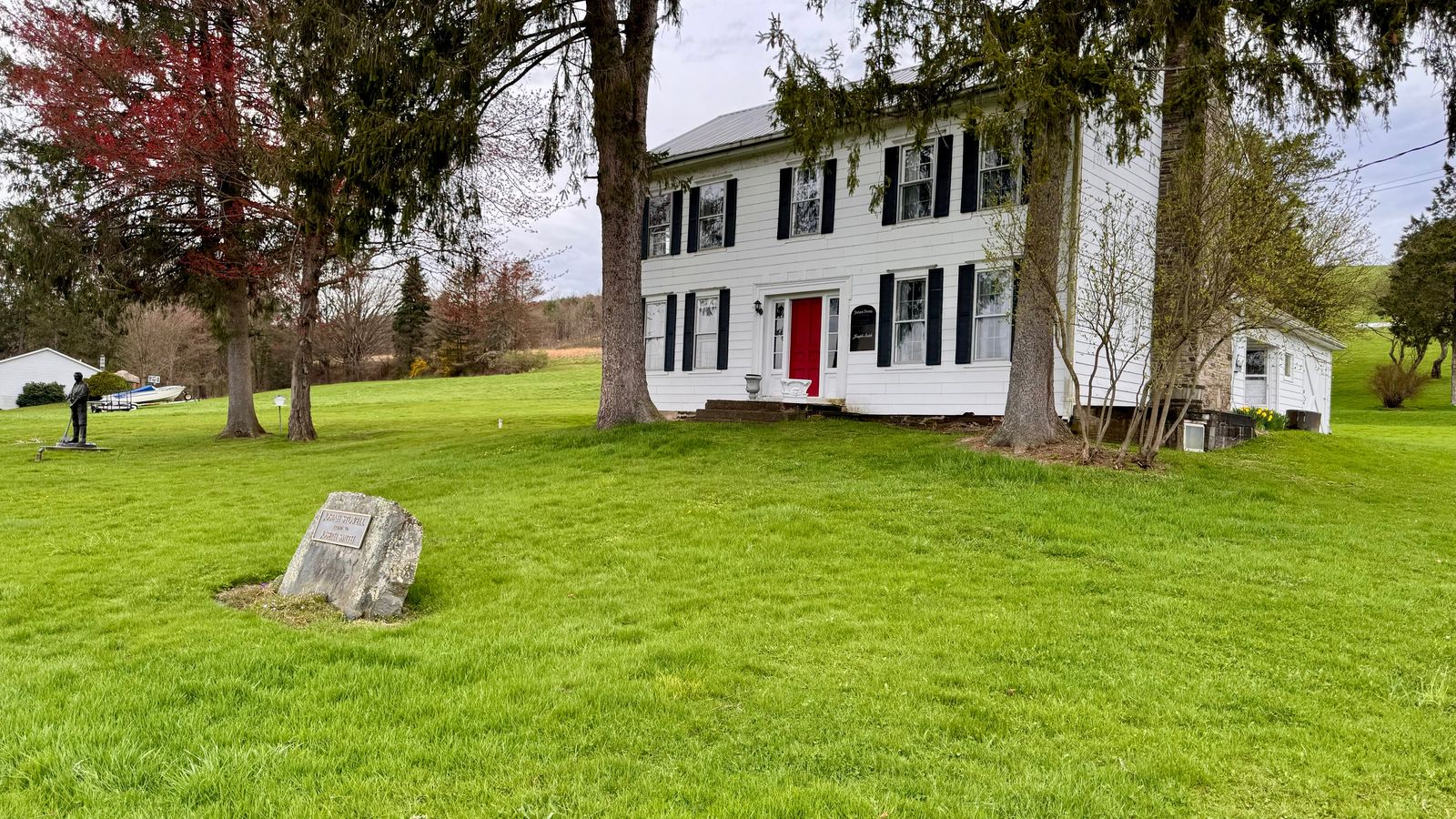 A two story white home with a red door surrounded by trees and a large stone in the front lawn.