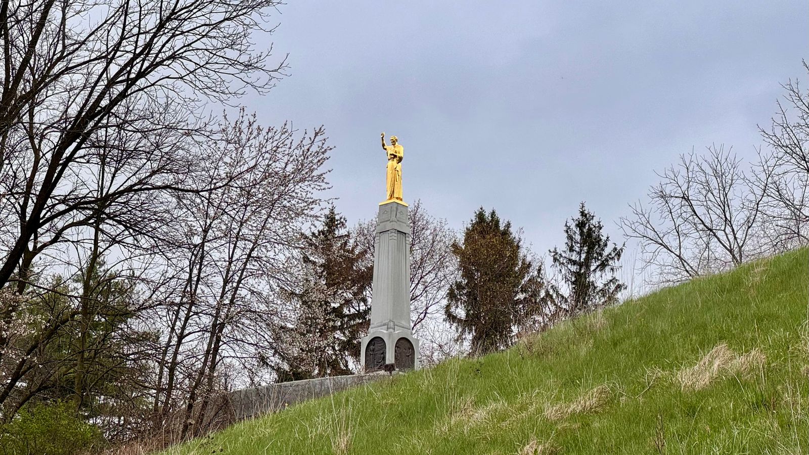 A tall stone piller with a large gold angel on top on a grassy hill.