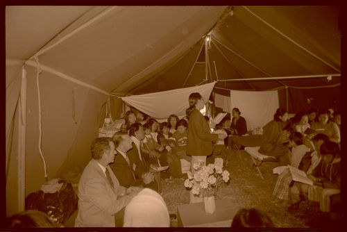 A Vietnamese woman stands at the center of an army base tent and leads a congregation to sing.