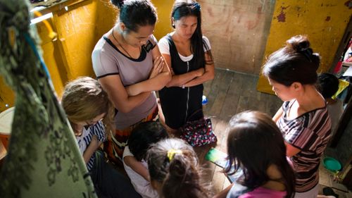 Three young sister missionaries in the Philippines.  They are shown walking on a dirt road by tin shack homes, teaching a family and praying with them in their home and as passengers on a motorcycle.