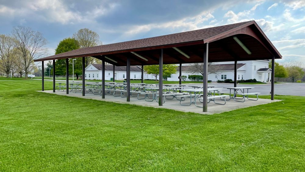 A pavilion with multiple metal picnic tables on a concrete slab in a grassy area by a road and building.