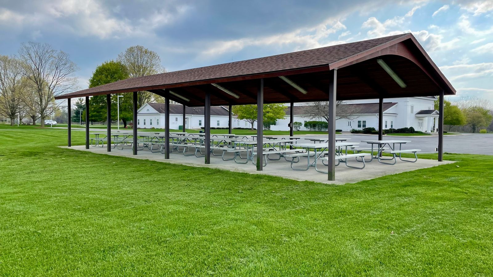 A pavilion with multiple metal picnic tables on a concrete slab in a grassy area by a road and building.