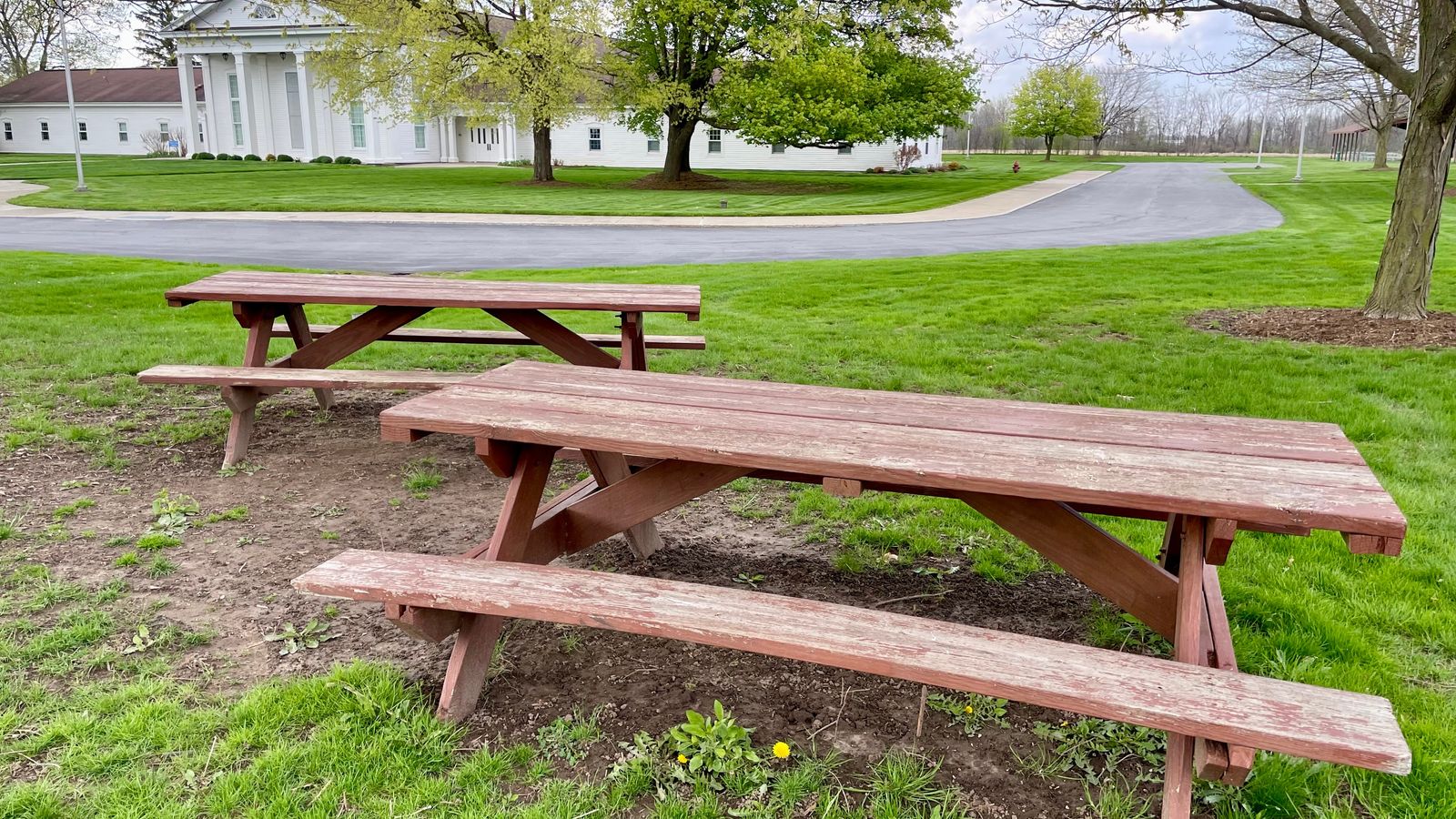 Two wooden picnic benches in a grassy area in front of a road and building. 