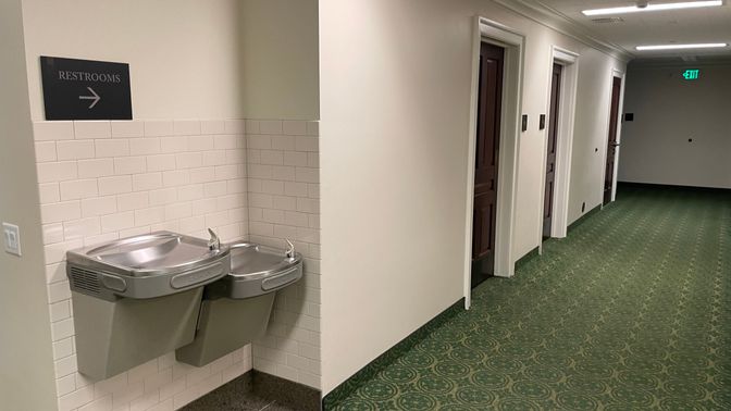 A hallway with green capet and water fountains on the left wall. Above the water fountains is a sign that says restrooms and farther down the hall are three wooden doors.