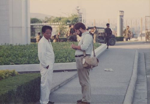 Dignardino Espi, dressed in a white jumpsuit, looks at the camera while a man takes notes and soldiers and military vehicles investigate near the temple fence.