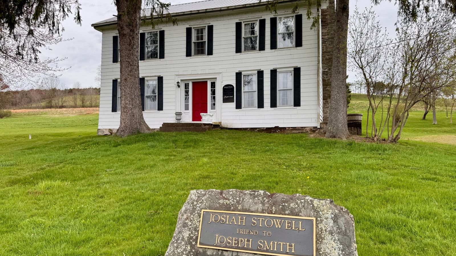 A two story white home with a red door surrounded by trees and a large stone with a metal plaque that reads Josiah Stowell friend to Joseph Smith.