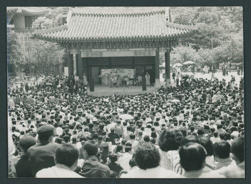 A crowd of hundreds surrounds a traditional Chinese stage where young men stand with guitars, a keyboard, and two clowns. A sign behind them reads “New Horizon” and “Tender Apples.”