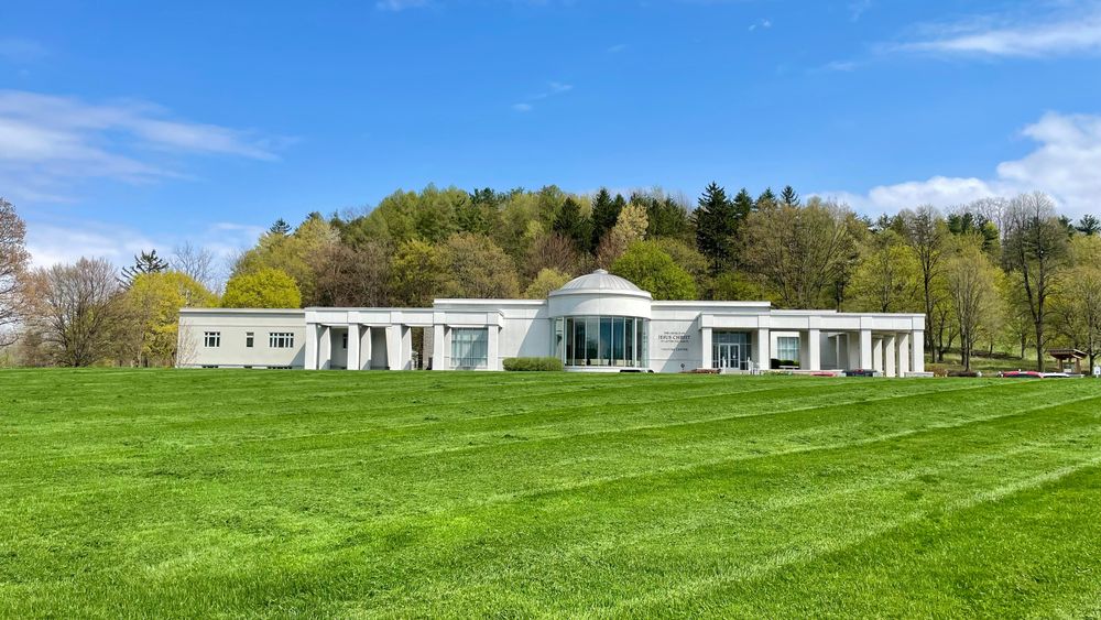 A long white building with a glass rotunda in the middle ontop of a grassy hill.