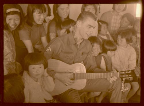 Stan Bronson, in uniform, sits and strums a guitar while a crowd girls looks on.