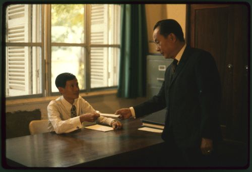 Nguyen Van The sits behind a desk in an office while another man hands him a tithing donation.